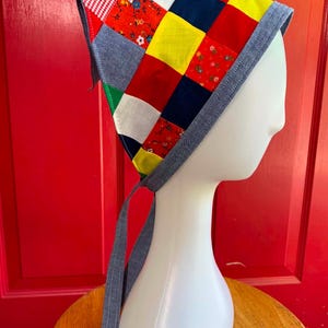 May include: A triangular headscarf featuring a patchwork design of multicolored fabric squares in red, yellow, blue, white, and green, including floral and gingham patterns. The headscarf is finished with a solid blue denim-like fabric border and ties.