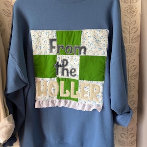 May include: A blue crewneck sweatshirt featuring a large, square, patchwork-style appliqué on the front. The appliqué consists of green and blue floral fabric squares with the text "From the HOLLER" stitched in dark gray and cream fabric letters.