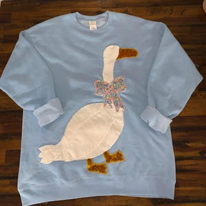 May include: A light blue sweatshirt featuring a large appliqué of a goose. The goose has a white body with corduroy texture, a brown beak and feet, and a floral bow tie. The sleeves are cuffed.