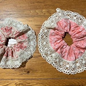 May include: Two handmade scrunchies. Each scrunchie features a pink floral fabric center, surrounded by a cream-colored lace trim. The scrunchies are displayed on a wooden surface.