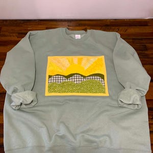 May include: A sage green crewneck sweatshirt featuring a rectangular fabric appliqué on the chest. The appliqué depicts a yellow sun rising over rolling hills, crafted from various patterned fabrics in shades of yellow, green, and grey gingham.