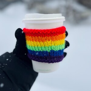 May include: A white disposable coffee cup with a rainbow-striped knitted cosy. The cosy features bands of red, orange, yellow, green, blue, and purple. The cup is held by a person wearing a black glove, with a snowy background.