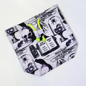 Pode incluir: Uma bolsa branca com cordão com um padrão em preto e branco com caveiras, aranhas, cobras e frascos de poções. A bolsa tem um cordão verde néon e o texto "HOUSE OF SALEM'S OLD CURIOSITY SHOP".