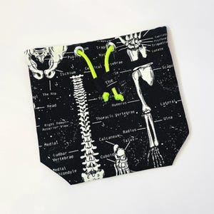 Pode incluir: Saco preto com cordão com uma impressão de anatomia esquelética branca e etiquetas ósseas. O saco tem um fecho de cordão verde néon. O design inclui a coluna vertebral, a anca e os ossos do braço.