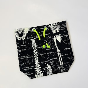 Pode incluir: Saco preto com cordão com um diagrama esquelético branco detalhado sobre um fundo preto. O saco inclui cordões e ilhós verde neon. Termos anatómicos como "Húmero" e "Vertebrae" são visíveis. Cerca de 20 cm de largura.