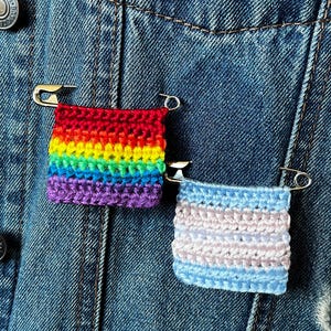 May include: Two crocheted pins pinned to a denim jacket. One pin has a rainbow design, and the other displays the colours of the transgender flag: light blue, pink, and white. The pins are secured with silver safety pins.