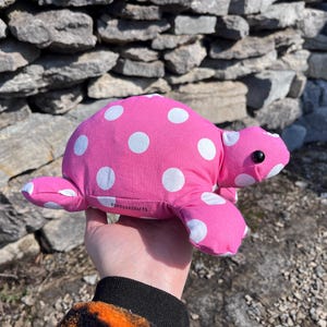 May include: A pink turtle-shaped soft toy with white polka dots. The toy has a black eye and four flippers. The turtle is made of fabric and is a cute, whimsical toy.
