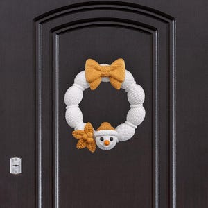 Könnte beinhalten: Ein weißer, gestrickter Schneemann-Kranz mit einer senffarbenen Schleife und Sternakzenten sowie einem Schneemann-Gesicht mit einer orangefarbenen Karottennase. Der Kranz hängt an einer dunkelbraunen Tür.