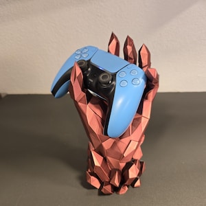 Puede incluir: Un mando de videojuegos azul claro y negro descansa en un soporte geométrico en forma de mano, de color marrón rojizo. El soporte tiene un diseño facetado, similar al cristal, que proporciona una exhibición única.