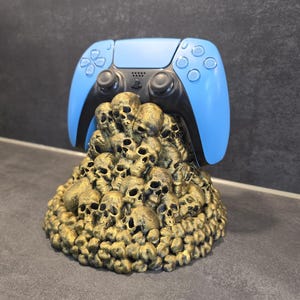 Peut inclure: Une manette de jeu bleue et noire repose sur une base dorée sur le thème des crânes. La manette a une face bleu clair et des boutons noirs. La base est une sculpture détaillée de nombreux crânes.