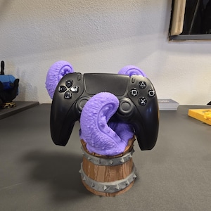 Könnte beinhalten: Ein schwarzer Videospiel-Controller ruht in einem holzfassförmigen Halter mit lila Tentakeln. Der Halter hat silberne Akzente und ein stacheliges Design um die Basis. Der Controller hat das Standard-Tastenlayout.