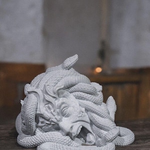 Op de afbeelding: Een grijze Medusa-sculptuur met slangen die zich om haar hoofd en gezicht wikkelen. De sculptuur heeft een open mond en gedetailleerde gelaatstrekken. Het kunstwerk is gemaakt van een steenachtig materiaal en staat op een houten oppervlak.