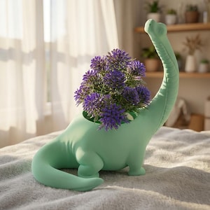Könnte beinhalten: Ein helles, minzgrünes Pflanzgefäß in Dinosaurierform mit langem Hals und geschwungenem Schwanz. Der hohle Rücken des Dinosauriers enthält ein Büschel leuchtend violetter Blumen mit texturierten Blütenblättern, die einen Kontrast zur glatten, matten Oberfläche bilden.