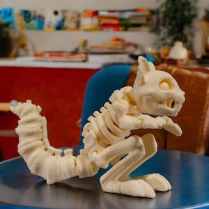 Könnte beinhalten: Eine cremefarbene, 3D-gedruckte Skulptur eines Cartoon-Kreaturen-Skeletts. Die Skulptur ist in einer stehenden Position mit eingerolltem Schwanz dargestellt. Die Figur steht auf einer blau-silbernen Oberfläche.