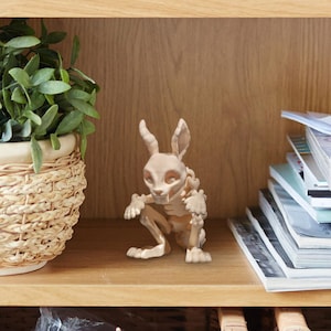 Könnte beinhalten: Beige Kaninchenskelett-Figur mit detaillierter Knochenstruktur. Die dekorative Figur sitzt auf einem Holzregal neben einer Topfpflanze und einem Stapel Zeitschriften. Ein einzigartiges Dekorationsstück für das Zuhause.