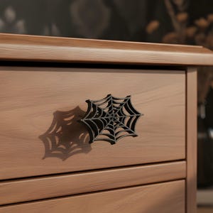 Könnte beinhalten: Ein schwarzer, spinnennetzförmiger Schubladengriff an einer hellbraunen Holzschubladenfront. Der Griff weist ein detailliertes, komplexes Netzmuster mit strahlenförmigen Linien und konzentrischen Kreisen auf, das einen Schatten wirft.