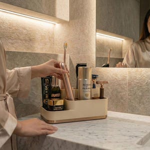 Könnte beinhalten: Ein beigefarbener und goldfarbener Badezimmer-Organizer mit Toilettenartikeln, darunter eine Zahnbürste, Zahnpasta und Hautpflegeprodukte. Der Organizer steht auf einer Marmorarbeitsplatte, mit einem Spiegel. Der Stil ist modern und minimalistisch.