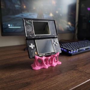 Könnte beinhalten: Eine schwarze tragbare Spielkonsole mit weißen grafischen Mustern auf dem Gehäuse, ausgestattet mit einem Dual-Screen-Design und einem Steuerkreuz. Die Konsole wird von einem leuchtend pinken, organisch geformten und strukturierten Kunststoffständer gehalten.