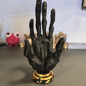 Könnte beinhalten: Eine schwarze, 3D-gedruckte Handskulptur mit detaillierter Textur, montiert auf einem Sockel mit gold-schwarzen Gefahrenstreifen. Die Hand befindet sich in einer offenen Position und zeigt die Finger und die Handfläche. Ein einzigartiger Dekorationsartikel.