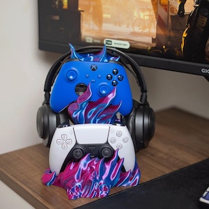 Può includere: Un supporto per accessori da gioco con una base a forma di fiamma blu e magenta che sostiene un controller wireless blu, un controller wireless bianco e un paio di cuffie nere. Il supporto è posizionato davanti a un monitor.