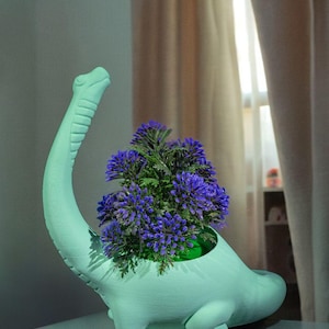 Può includere: Un vaso a forma di dinosauro verde menta con fiori viola. Il vaso ha un lungo collo e un corpo arrotondato, con i fiori che riempiono l'apertura. Il vaso è su una superficie azzurra.