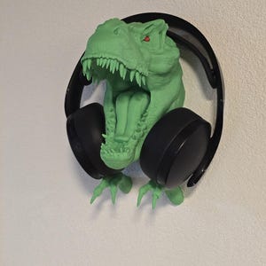 Könnte beinhalten: Ein grüner Kopfhörerhalter in Form eines Tyrannosaurus Rex mit roten Augen und scharfen Zähnen. Schwarze Kopfhörer sind im Maul des Dinosauriers platziert. Der Halter ist an einer weißen Wand befestigt.