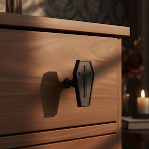 Könnte beinhalten: Ein mattschwarzer Schubladengriff in Sargform mit einem erhabenen Kreuzdetail auf der Vorderseite. Der Griff ist an einer hellbraunen Holzschubladenfront mit sichtbarer horizontaler Holzmaserung und einer glatten Oberfläche befestigt.