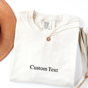 May include: Cream-colored t-shirt with the text "Custom Text" in black. A gold necklace with a pendant rests on the shirt. A brown hat and dried leaves are partially visible in the background. The shirt has a Comfort Colors tag.