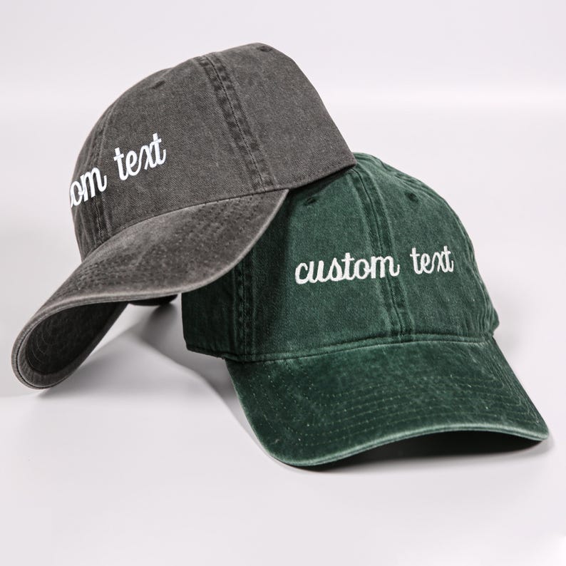 Puede incluir: Dos gorras de b&eacute;isbol ajustables con bordado de texto personalizado. Una gorra es gris oscuro y la otra es verde oscuro.