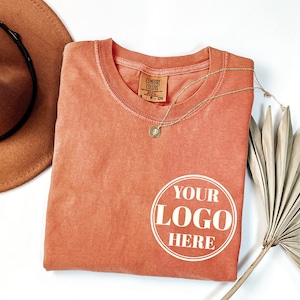 May include: A folded coral-colored t-shirt with the text "YOUR LOGO HERE" in a white circle on the front. The shirt is made by Comfort Colors.
