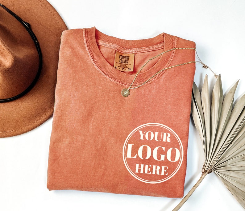 May include: A folded coral-colored t-shirt with the text "YOUR LOGO HERE" in a white circle on the front. The shirt is made by Comfort Colors.