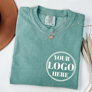 May include: A teal blue t-shirt with a white circle that says "YOUR LOGO HERE" on the front. The shirt is folded and laid flat on a white surface.