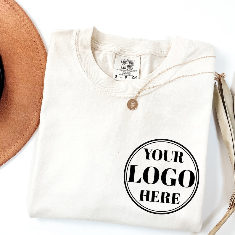 May include: A white t-shirt with a black circle that says "YOUR LOGO HERE" on it. The shirt is folded and has a tag that says "COMFORT COLORS" on it.