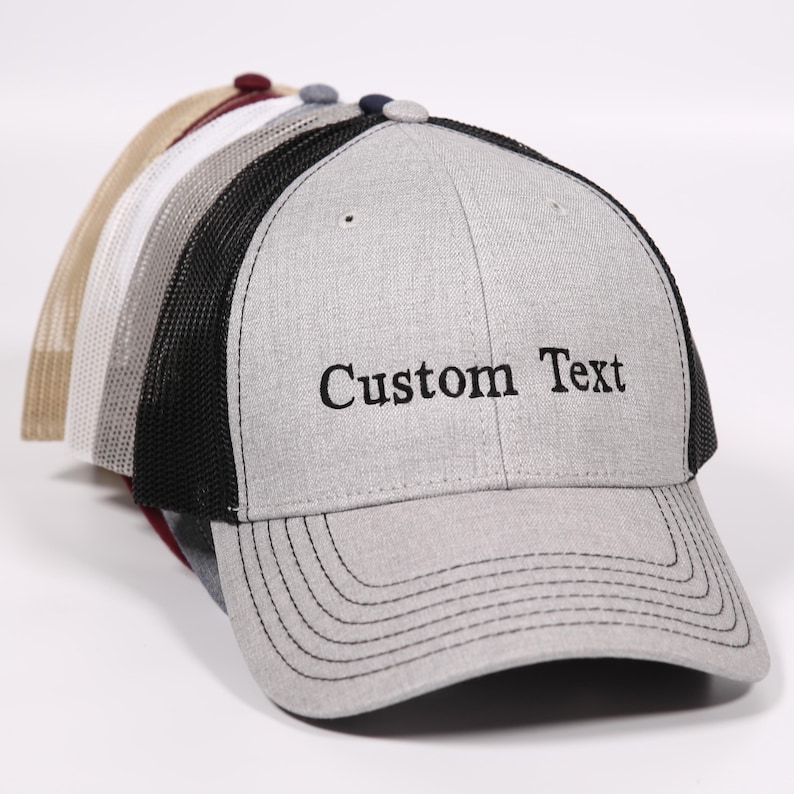 Puede incluir: Una gorra de camionero gris y negra con una parte trasera de malla negra. La gorra tiene una visera curvada y est&aacute; bordada con el texto "Custom Text".