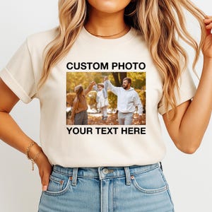 May include: A cream-colored t-shirt featuring a family photo with the text "CUSTOM PHOTO" above the image and "YOUR TEXT HERE" below. The photo shows a family of three holding hands in a park setting.
