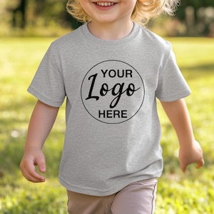 May include: A light grey t-shirt featuring a black circular logo with the text "YOUR Logo HERE". The short-sleeved shirt is made of a soft material. The t-shirt is being worn by a child.