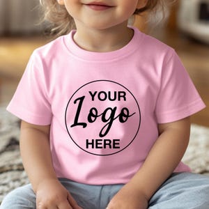 May include: A light pink t-shirt with a crew neck. The shirt features a black circular logo with the words "YOUR Logo HERE" in a handwritten style font. The shirt is made of a soft material.