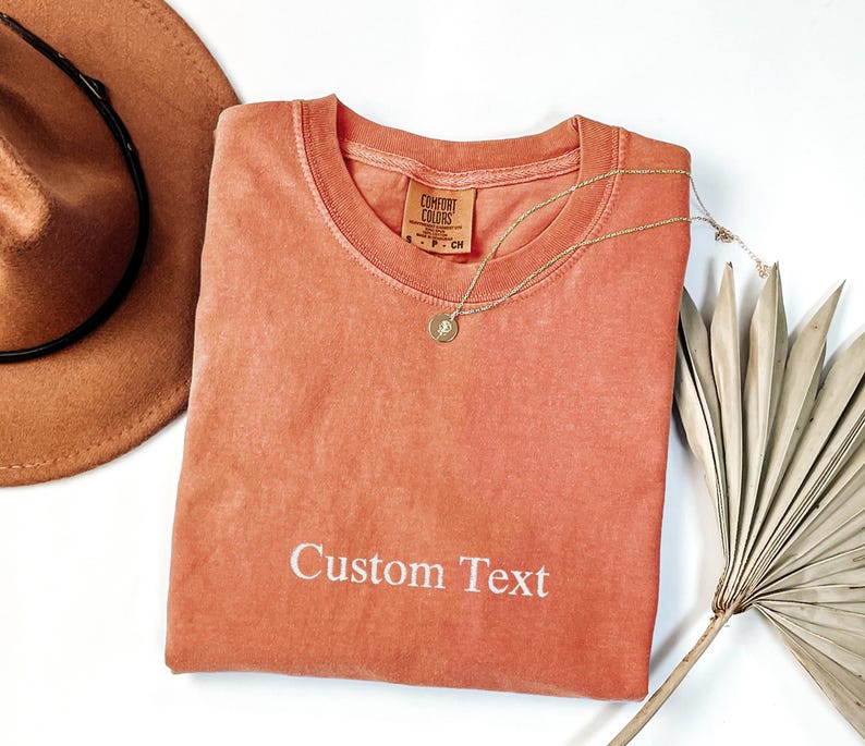 May include: A coral-colored t-shirt with the text "Custom Text" in white. The shirt has a round neckline and a small tag. A gold necklace with a pendant and a brown hat are also in the image.