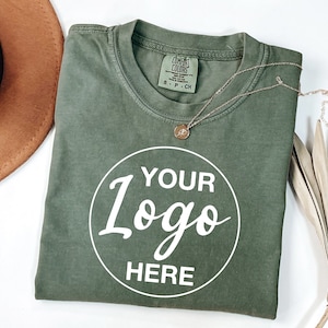 May include: A green t-shirt with a white circle that says "YOUR Logo HERE" on it. The shirt is folded and laid flat on a white surface. There is a brown hat and a gold necklace on the left side of the shirt and a dried palm leaf on the right side of the shirt.
