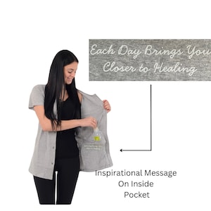 May include: A light gray short-sleeved button-down cardigan featuring an internal pocket. The inside of the pocket is printed with the white text "Each Day Brings You Closer to Healing." The garment is worn over a black top and black leggings.