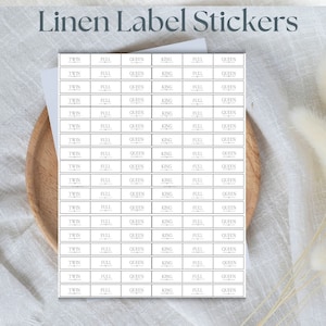 Op de afbeelding: Een vel witte linnen etiket stickers met de woorden "Twin", "Full", "Queen" en "King" gedrukt op elke sticker. De stickers zijn ontworpen om beddengoed maten te labelen.