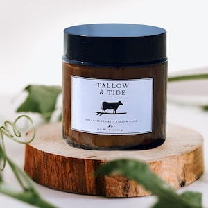 May include: A brown glass jar with a black lid, labeled "Tallow & Tide" with a silhouette of a cow surfing. The jar sits on a wooden slice and contains 4 oz (114 g) of balm. The background is white with green foliage.