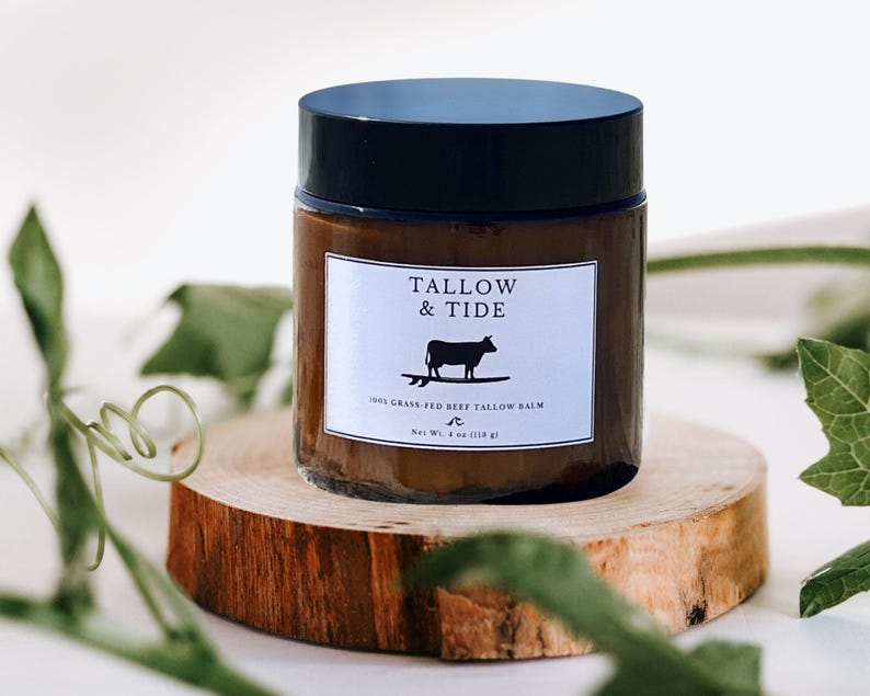 May include: A brown glass jar with a black lid, labeled "Tallow & Tide" with a silhouette of a cow surfing. The jar sits on a wooden slice and contains 4 oz (114 g) of balm. The background is white with green foliage.