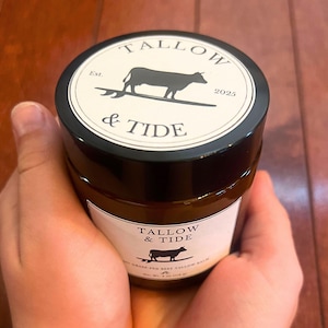 May include: A jar of Tallow & Tide beef tallow balm held in hands. The jar has a black lid with a white label featuring a cow silhouette and the text "Tallow & Tide". The jar's label also includes the text "Est. 2025".