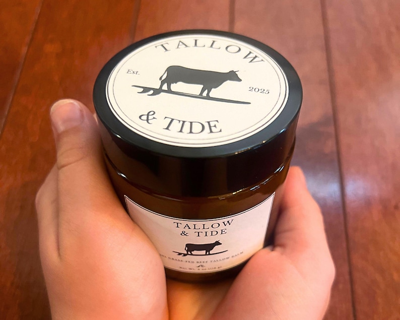 May include: A jar of Tallow & Tide beef tallow balm held in hands. The jar has a black lid with a white label featuring a cow silhouette and the text "Tallow & Tide". The jar's label also includes the text "Est. 2025".