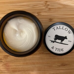 May include: An open, dark amber jar filled with a white, creamy substance next to a black lid. The lid features the text "TALLOW & TIDE" around a black silhouette of a cow on a surfboard, with "Est. 2025" below.