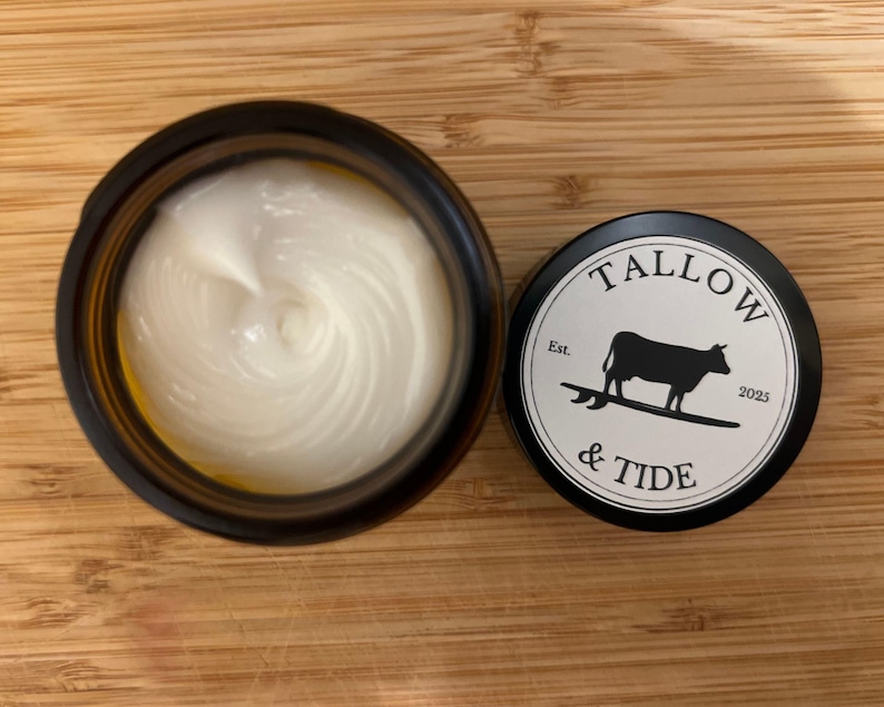 May include: An open, dark amber jar filled with a white, creamy substance next to a black lid. The lid features the text "TALLOW & TIDE" around a black silhouette of a cow on a surfboard, with "Est. 2025" below.