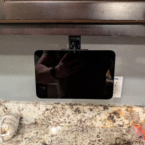 Puede incluir: Una tableta negra está montada debajo de un armario marrón. La tableta se sujeta con un soporte negro. El fondo incluye una pared de color claro, una encimera y una toma de corriente.