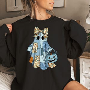 May include: Black sweatshirt featuring a whimsical Halloween ghost design. The ghost is patchwork-style, with a blue body, a bow, and a pumpkin basket. The design incorporates floral and patterned fabric elements, perfect for the spooky season.