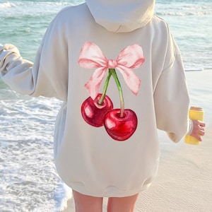 May include: A light gray hoodie with a pink bow and two red cherries on the back. The cherries have green stems and leaves.